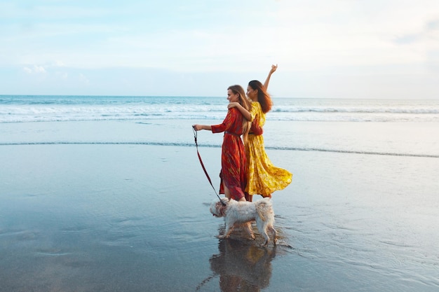 Due donne in abiti maxi che camminano con il cane lungo la riva dell'oceano