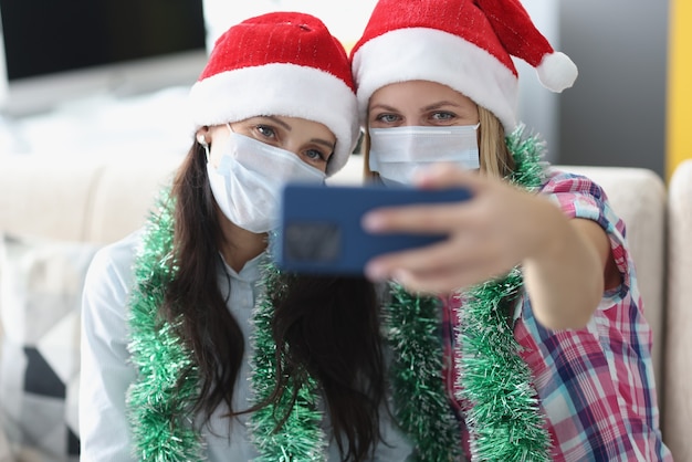 Due donne in cappelli di Babbo Natale e maschere protettive mediche sono fotografate sul ritratto della fotocamera del telefono