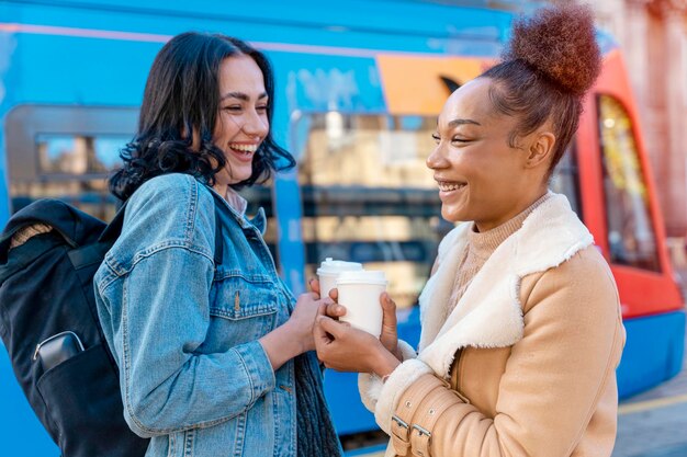 Due donne in giacca di jeans parlano bevendo caffè e aspettano il tram alla fermata Foto di stile di vita