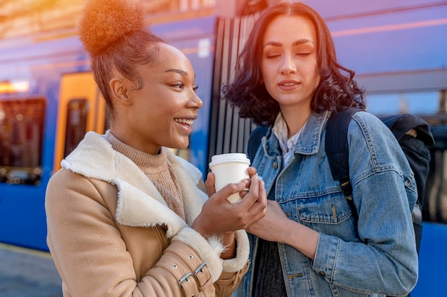 Due donne in giacche di jeans parlano tra loro ridendo bevendo caffè e aspettando un tram