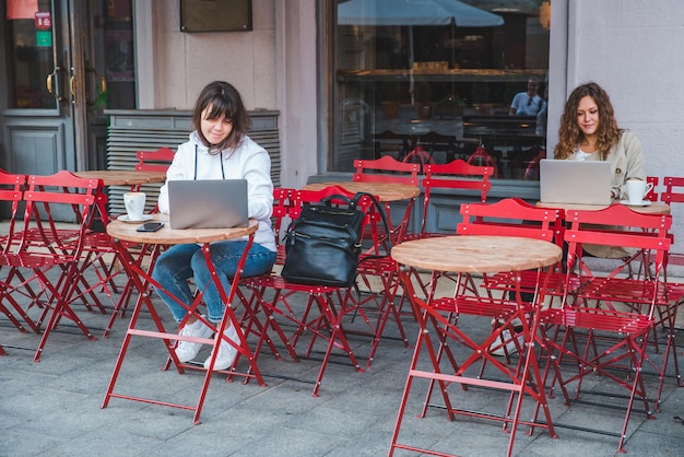 Due donne in un caffè seduto a un tavolo diverso che beve latte lavorando sul computer portatile