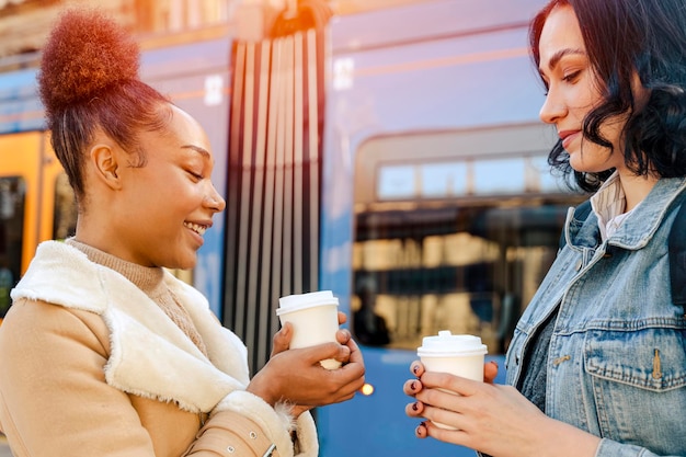 Due donne in una giacca di jeans parlano tra loro ridendo bevendo caffè e aspettando un tram