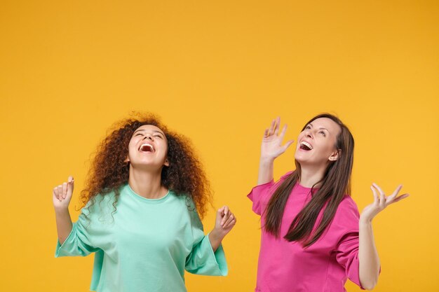 Due donne ridenti amiche ragazze afroamericane europee in abiti rosa verdi in posa isolata su sfondo giallo arancio. Concetto di stile di vita delle persone. Mock up spazio di copia. Alzando lo sguardo, allargando le mani.