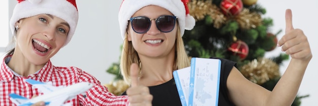 Due donne sorridenti in cappelli di Babbo Natale tengono i biglietti e salutano la vacanza e il viaggio