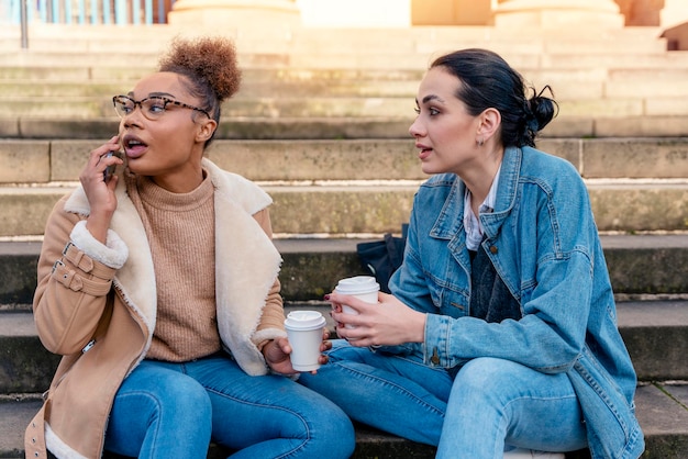 Due donne studentesse in giacca di jeans che parlano tra loro al telefono ridendo bevendo caffè sulle scale