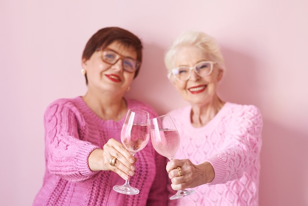 due eleganti donne anziane in maglioni rosa con bicchieri di vino rosato su sfondo rosa