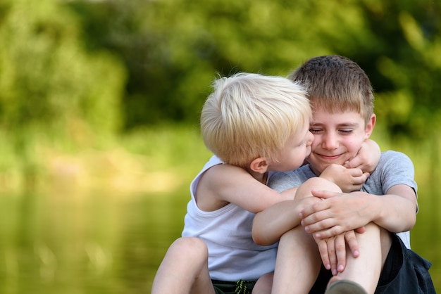 Due fratelli piccoli sono seduti all'aperto. Uno bacia l'altra guancia. Alberi verdi sfocati in lontananza. amicizia e fratellanza.