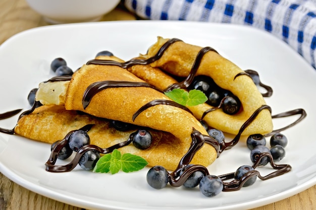 Due frittelle con mirtilli, menta e glassa di cioccolato su un piatto bianco, tovagliolo, ciotola sullo sfondo di tavole di legno