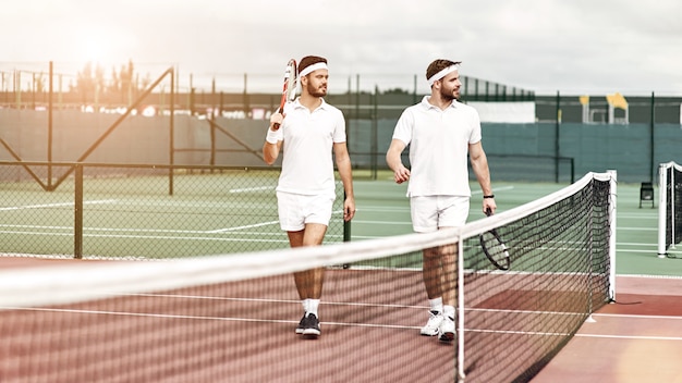 Due giocatori di tennis in abiti sportivi bianchi con le racchette in mano che camminano sul campo