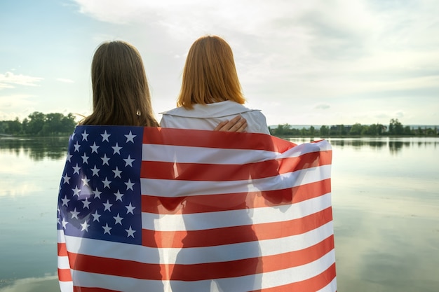 Due giovani amiche donne con la bandiera nazionale degli Stati Uniti sulle spalle che si abbracciano insieme