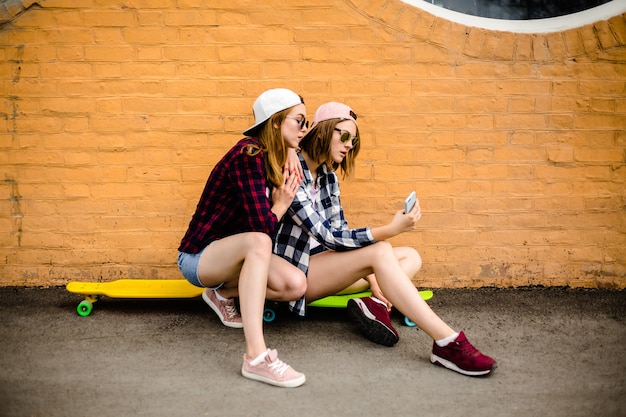 Due giovani amiche felici in abito hipster seduti su longboard e facendo selfie sul telefono.