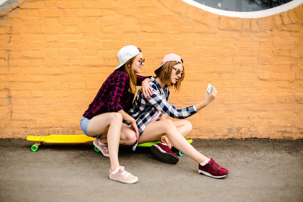 Due giovani amiche felici in abito hipster seduti su longboard e facendo selfie sul telefono.