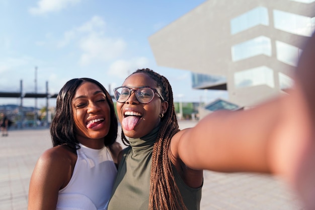 Due giovani amiche nere si divertono a tirare fuori la lingua per scattare una foto selfie con un telefono cellulare, concetto di giovinezza e amicizia, copia spazio per il testo