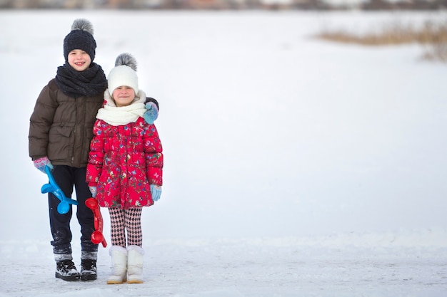 Due giovani bambini sorridenti felici svegli in vestiti caldi con nuove clip luminose della neve che posano insieme il giorno freddo di inverno sullo spazio vago luminoso bianco della copia. Attività all'aperto, giochi per le vacanze.