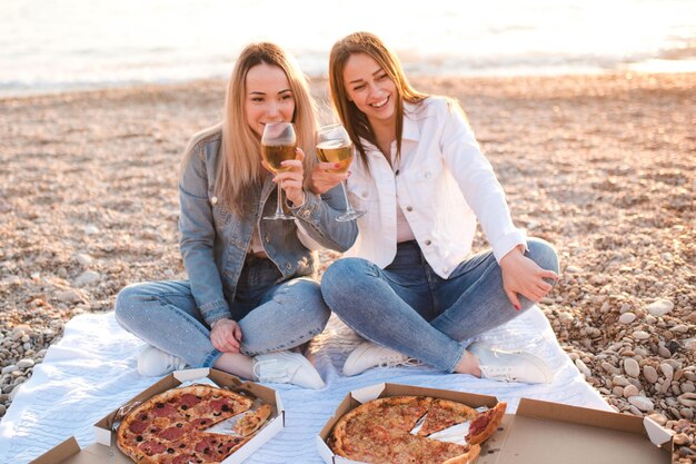 Due giovani belle amiche che si divertono con pizza e vino in spiaggia sulla riva del mare all'aperto alla luce del sole. Stagione delle vacanze estive. Amicizia. Felicità. Le sorelle trascorrono del tempo insieme sulla costa.