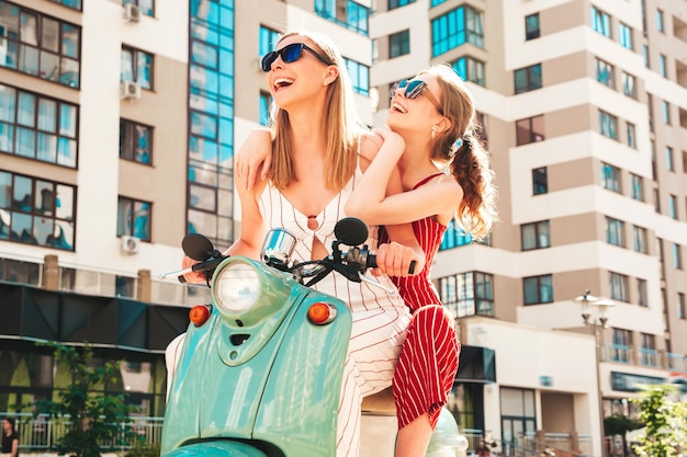 Due giovani belle donne hipster sorridenti in tuta alla modaDonne spensierate sexy che guidano una moto retrò sullo sfondo della stradaModelli positivi che si divertono a cavalcare un classico scooter italiano in occhiali