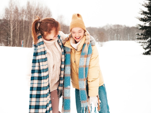 Due giovani belle donne sorridenti hipster in vestiti caldi alla moda e sciarpe. Donne spensierate in posa in strada nel parco. Modelli puri positivi che si divertono nella neve. Godersi i momenti invernali. concetto di natale