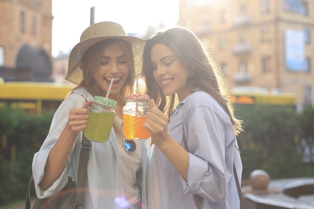 Due giovani belle donne sorridenti in abiti estivi si divertono in città e bevono cocktail freschi.