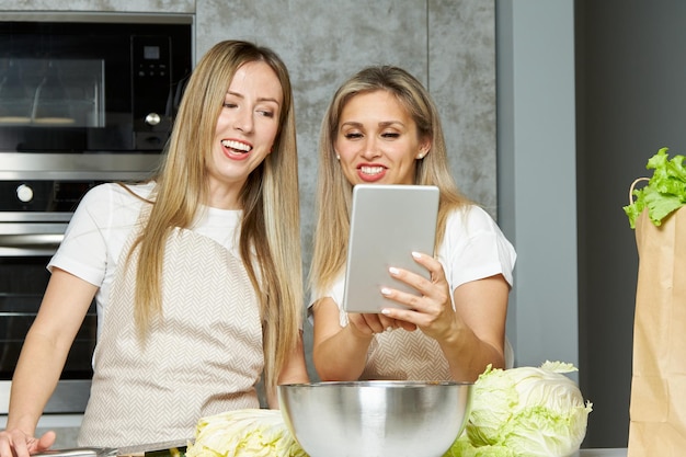 Due giovani belle donne usano un tablet digitale per bloggare mentre cucinano in cucina a casa amici sorridenti felici preparano cibo sano in cucina e trasmettono online usando il tablet