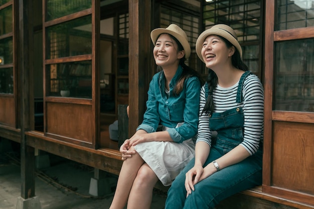 Due giovani belle ragazze in denim indossano e cappelli di paglia seduti sui gradini a parlare e ridere. amiche che si rilassano all'aperto a Tokyo in Giappone. le donne sorridenti godono della vista del giardino dalla vecchia casa di legno