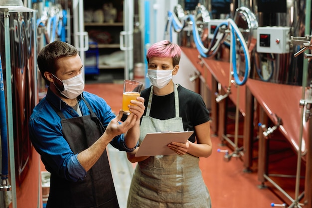 Due giovani birrai che guardano un bicchiere di birra fresca e discutono delle sue caratteristiche e del suo gusto mentre una donna li scrive in officina