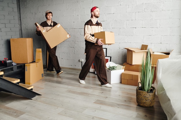 Due giovani caricatori in abiti da lavoro che trasportano scatole di cartone imballate mentre aiutano a consegnare pacchi a un nuovo appartamento, casa o studio