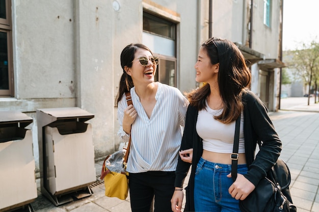 Due giovani donne allegre che ridono e che camminano all'aperto. gruppo di amiche coreane asiatiche che si rilassano all'ora del tramonto con gli occhiali da sole si guardano l'un l'altro. le sorelle si godono il sole sulla strada nella città vecchia