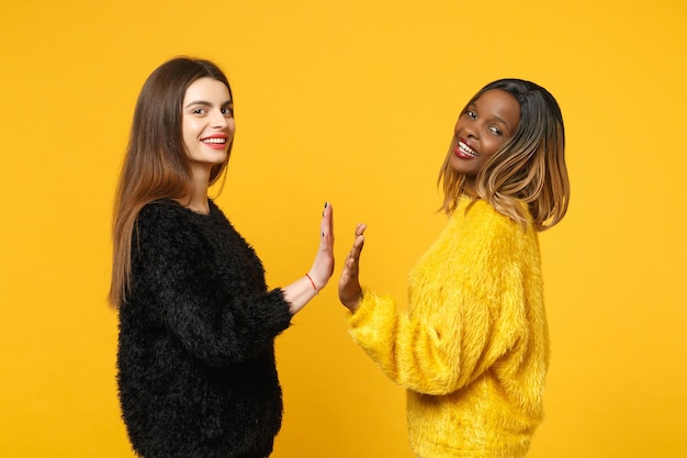 Due giovani donne amiche europee e afroamericane in abiti gialli neri in piedi in posa isolata su sfondo arancione brillante, ritratto in studio. Concetto di stile di vita delle persone. Mock up spazio di copia.