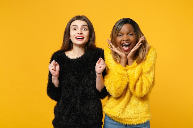 Due giovani donne amiche europee e afroamericane in abiti gialli neri in piedi in posa isolata su sfondo arancione brillante, ritratto in studio. Concetto di stile di vita delle persone. Mock up spazio di copia.