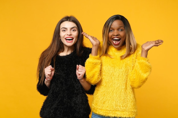 Due giovani donne amiche europee e afroamericane in abiti gialli neri in piedi in posa isolata su sfondo arancione brillante, ritratto in studio. Concetto di stile di vita delle persone. Mock up spazio di copia.