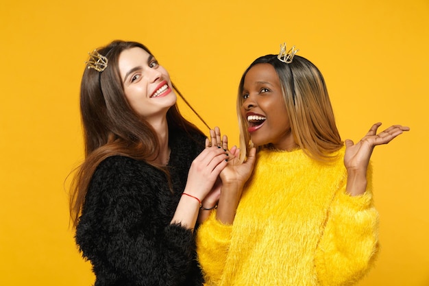 Due giovani donne amiche europee e afroamericane in abiti gialli neri in piedi in posa isolata su uno sfondo di parete arancione brillante, ritratto in studio. Concetto di stile di vita delle persone. Mock up spazio di copia.