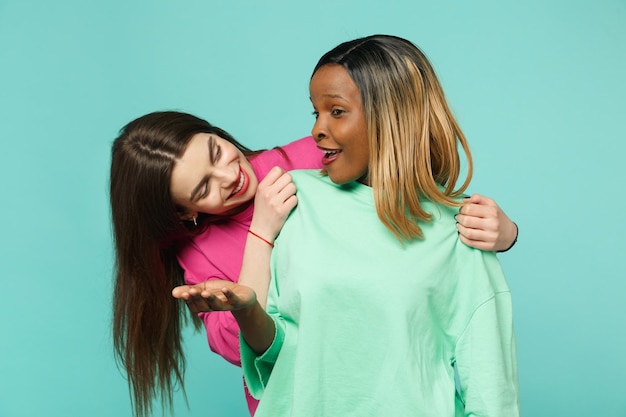 Due giovani donne amiche europee e afroamericane in abiti rosa verdi in piedi in posa isolata su sfondo blu turchese muro, ritratto in studio. Concetto di stile di vita delle persone. Mock up spazio di copia.