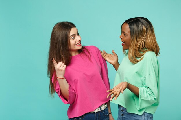 Due giovani donne amiche europee e afroamericane in abiti rosa verdi in piedi in posa isolata su sfondo blu turchese muro, ritratto in studio. Concetto di stile di vita delle persone. Mock up spazio di copia.