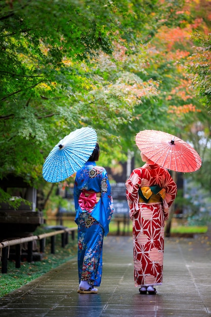 Due giovani donne asiatiche che indossano kimono blu e rosso con ombrello tenendo la mano a piedi nel parco alla stagione delle foglie d'autunno in giappone