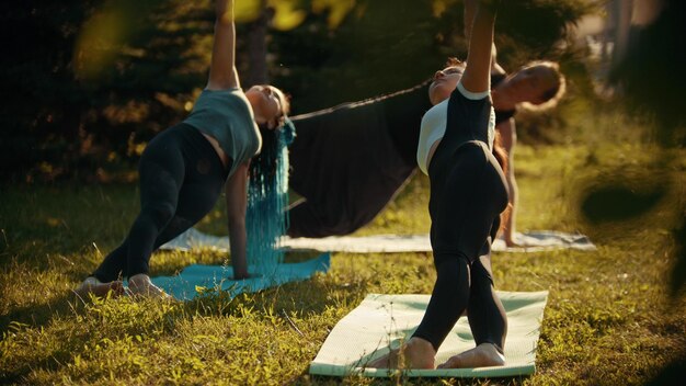 Due giovani donne atletiche e un uomo che fanno esercizi di yoga nel parco circondato da alberi verde brillante