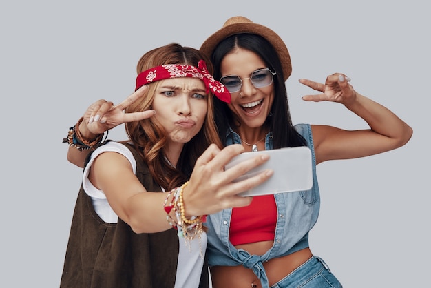 Due giovani donne attraenti ed eleganti che si fanno selfie e sorridono mentre sono in piedi su sfondo grigio