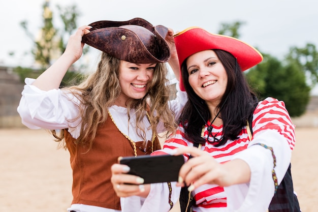 Due giovani donne caucasiche sorridenti felici in costumi del pirata che prendono selfie sullo smartphone.