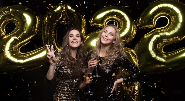 Due giovani donne che bevono champagne. Immagine di ragazze con palloncini isolati su sfondo nero, divertirsi alla festa di Natale o Capodanno 2022.