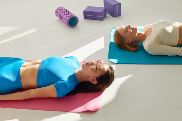 Due giovani donne che fanno la postura Savasana dopo la lezione di yoga Concetto di cura della salute fisica e mentale meditazione profonda Etnia asiatica e caucasica