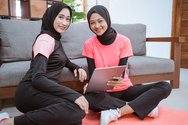 Due giovani donne che indossano abbigliamento sportivo hijab sorridente mentre ci si rilassa sul pavimento mentre si utilizza una tavoletta digitale appoggiata sul divano di casa