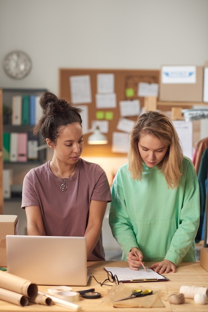 Due giovani donne che lavorano in squadra al tavolo con computer e documenti in piedi in officina