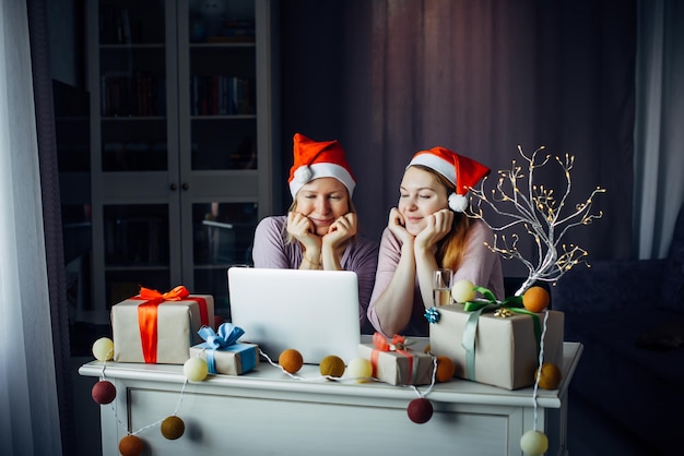 Due giovani donne con i cappelli di Babbo Natale sedute a tavola con il computer portatile tra ghirlande e regali e augurano buon Natale. Videochiamate a Capodanno 2022.