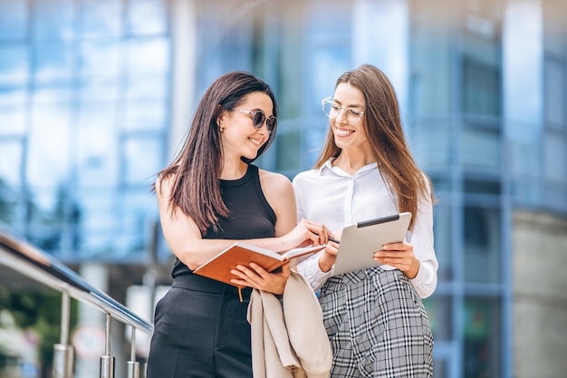 Due giovani donne d'affari con tablet e notebook che camminano all'aperto vicino al moderno centro business.