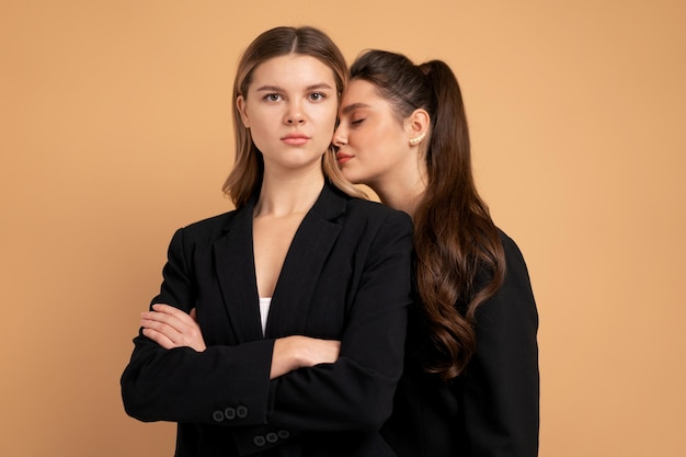 Due giovani donne d'affari vestite di nero in piedi in studio di colore arancione di sfondo. Una donna che guarda la telecamera con le braccia incrociate sul petto, un'altra donna le ha messo la testa sulla spalla. Supporto degli amici