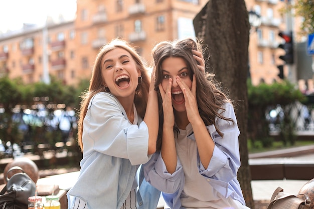 Due giovani donne hipster sorridenti in abiti estivi in posa sulla strada. Femmina che mostra emozioni positive per il viso.