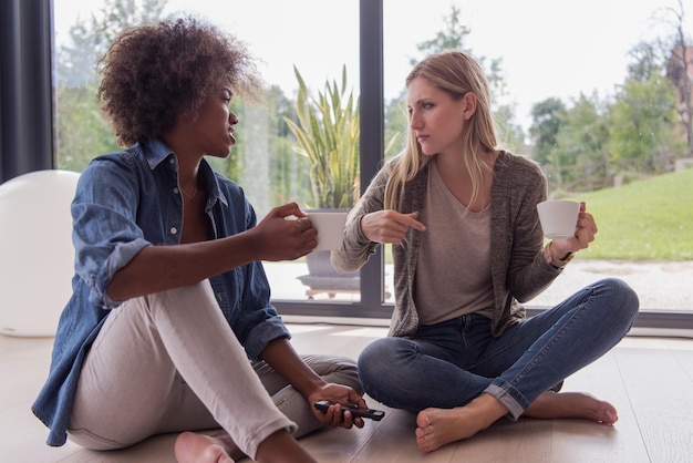 Due giovani donne multietniche che ridono si siedono sul pavimento vicino alla finestra e si divertono bevendo un caffè usando lo smartphone in una casa di lusso