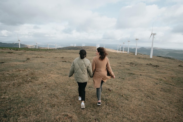 Due giovani donne, su panni invernali durante una giornata ventosa, si godono una giornata divertendosi a correre insieme tra il mulino a vento elettrico. Stile di vita. Stile hipster e moderno. Stile cinematografico.