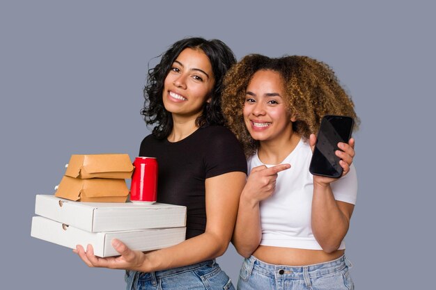 Due giovani donne, una latina e una con i capelli afro, ridono tenendo in mano pizze e hamburger
