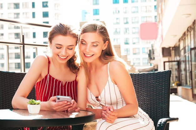 Due giovani e belle donne hipster sorridenti in tute estive alla moda Donne spensierate che chattano nel caffè della verandaLa modella mostra le informazioni della sua amica nel suo smartphone Guardano lo schermo e usano le app