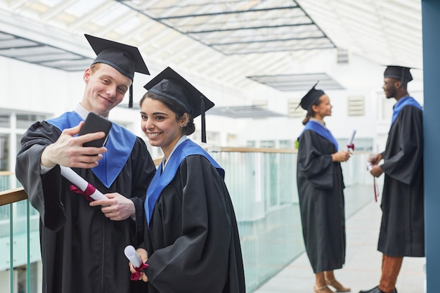 Due giovani felici che indossano abiti da laurea e tengono in mano uno smartphone mentre si fanno selfie in interni all'interno del college